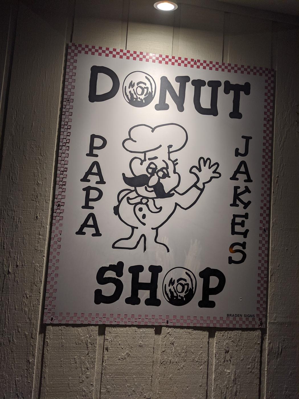 The painted Papa Jake's Donut Shop sign, black hand drawn lettering and a moustached baker on white inside a red and white checkerboard border.